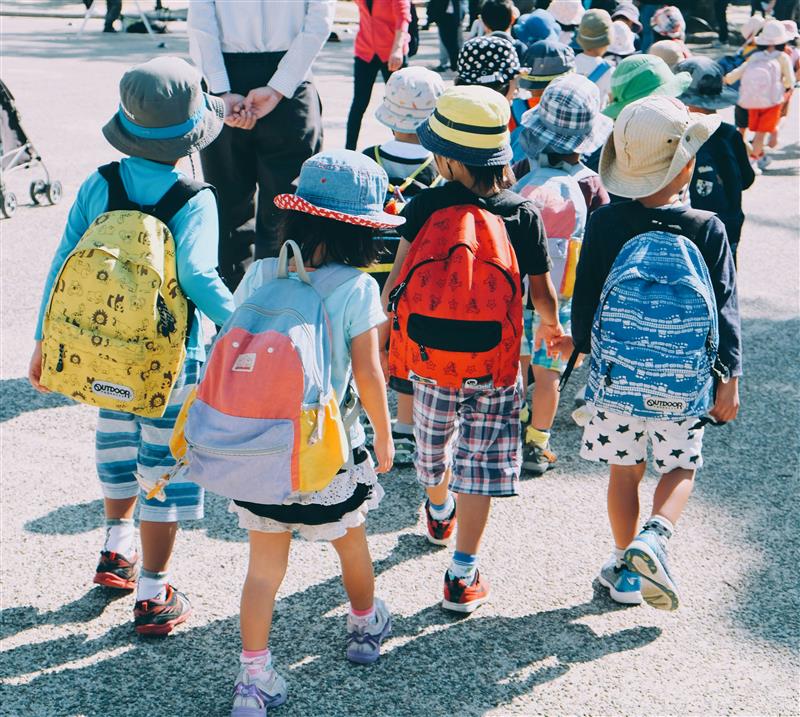 From the back, a group of children in hats take a walk in the sunshine.
