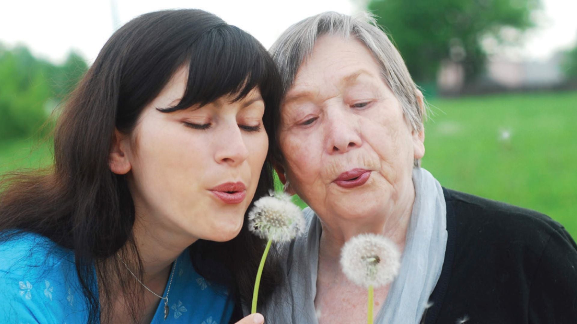 An older woman and a young woman blow wishes on dandelions.