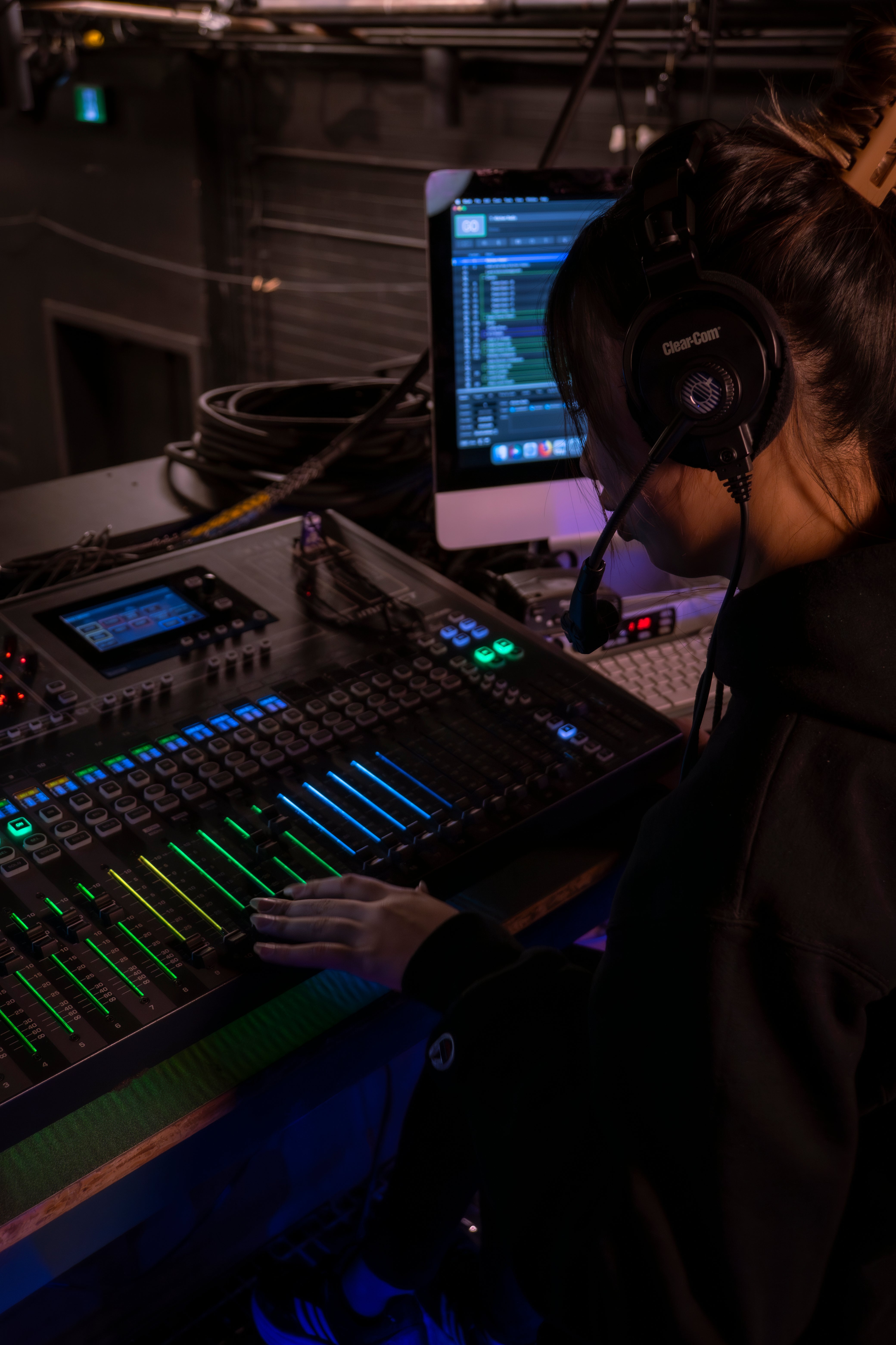 A person sit in front of a technical board glowing green and blue. They are wearing a headset and looking down intently at the board.