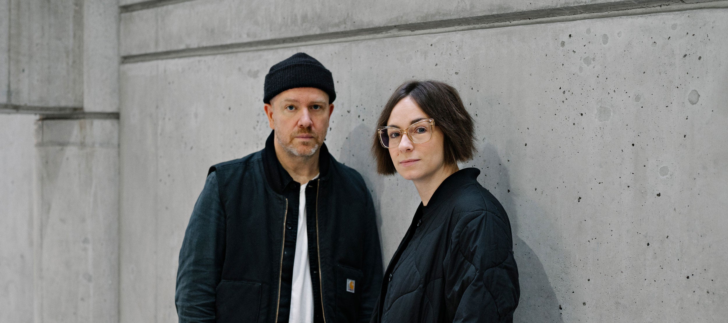 Tanya Goehring and Trevor Jacobson (The Automatic Message) posing against a grey wall.