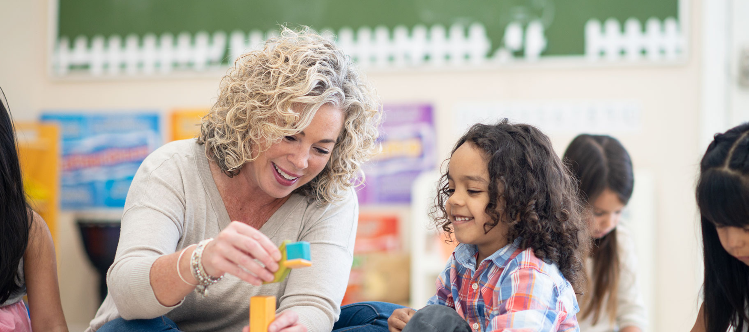Blonde woman playing blocks with a young student in a classroom setting.