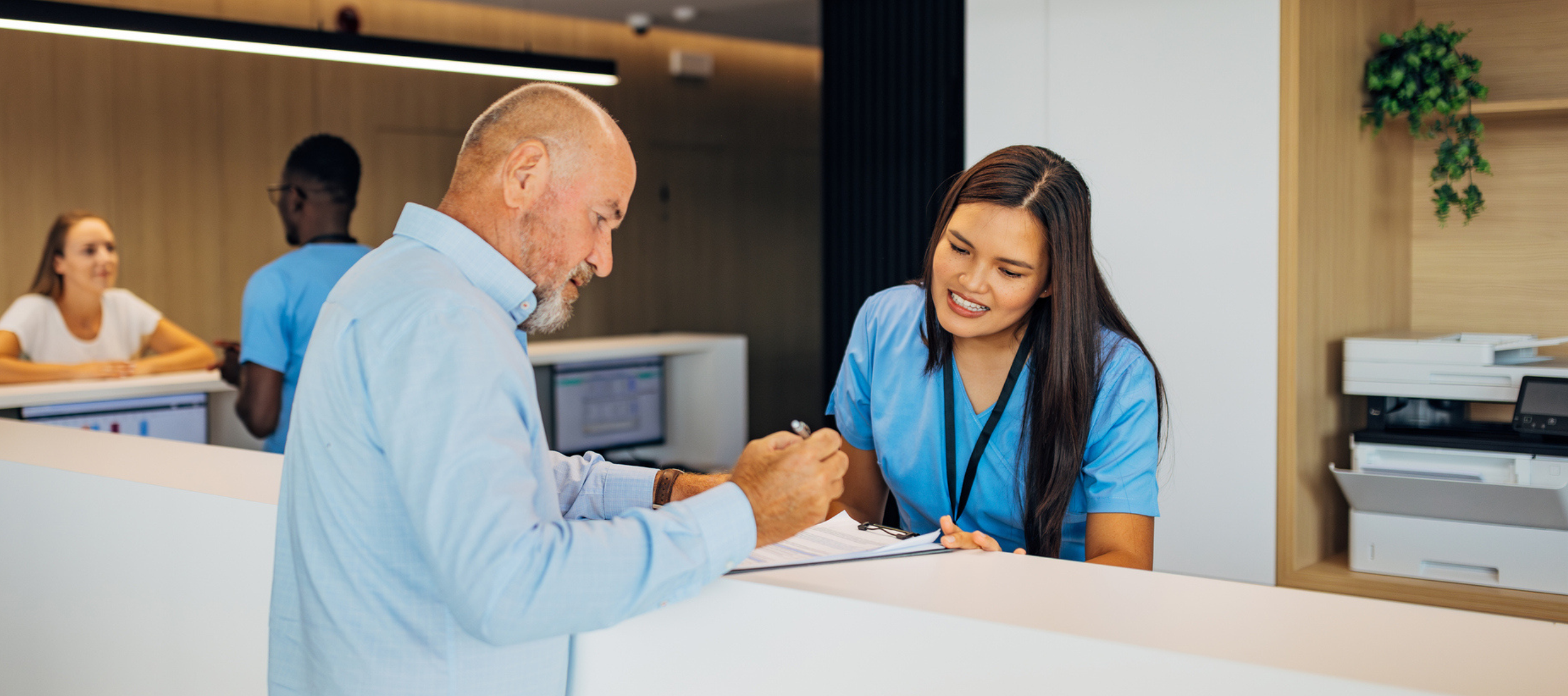 Stock image of the front desk at a medical office, with an office administrator assisting a patient.