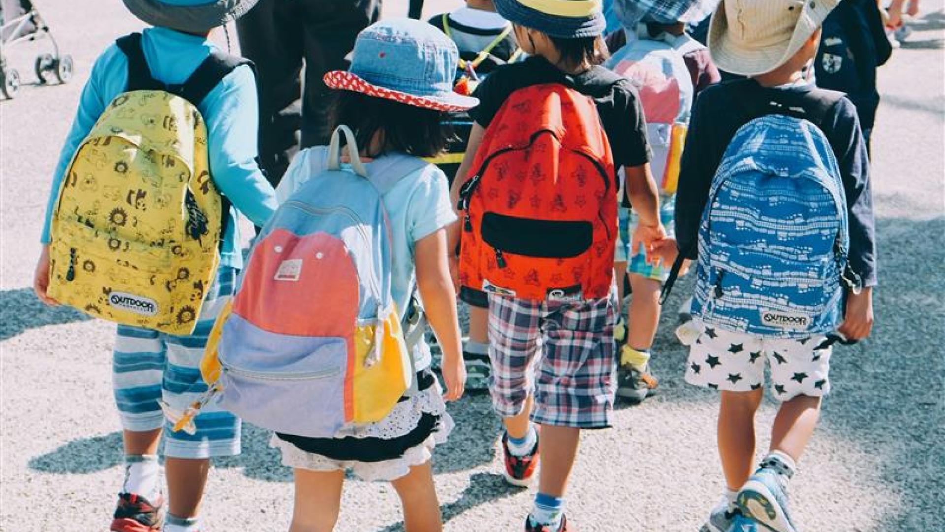 From the back, a group of children in hats take a walk in the sunshine.