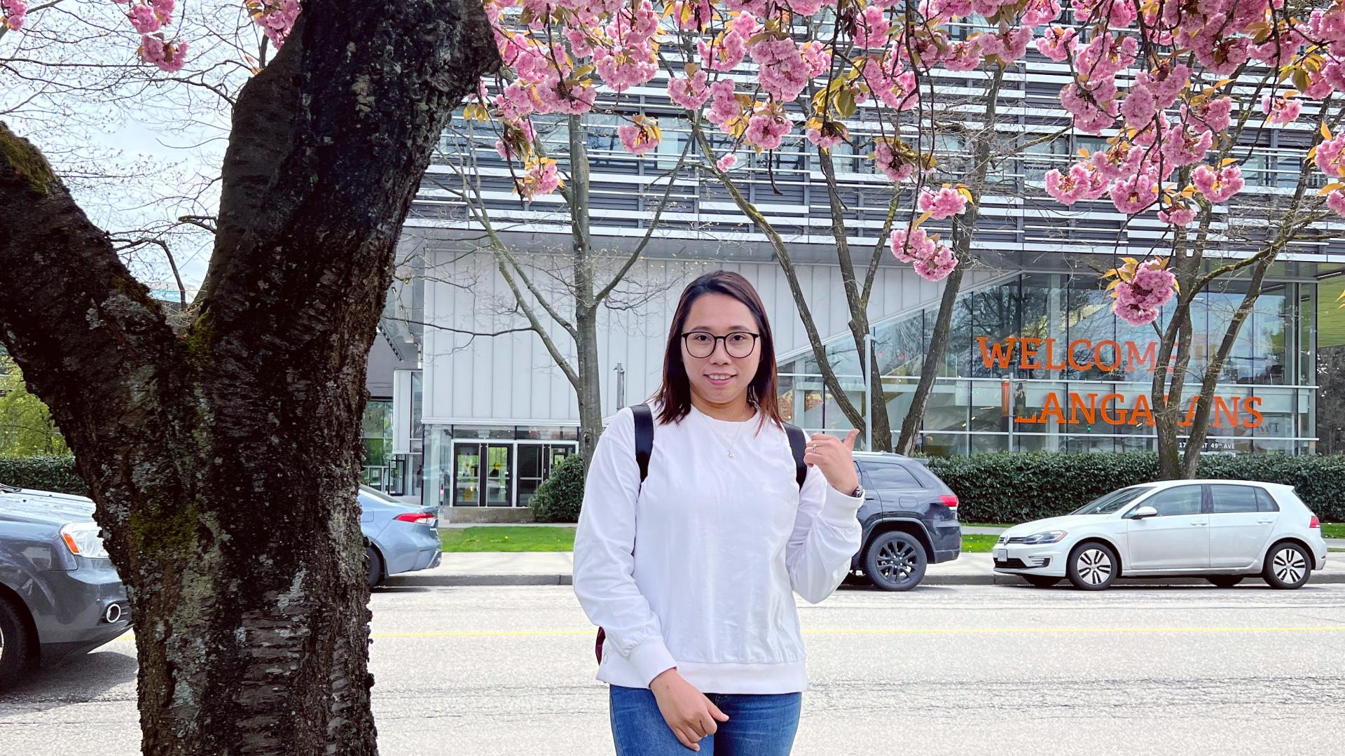 Hnin (Elisa) Pwint under a cherry blossom tree across the street from Langara College with the Science and Technology Building in the background.