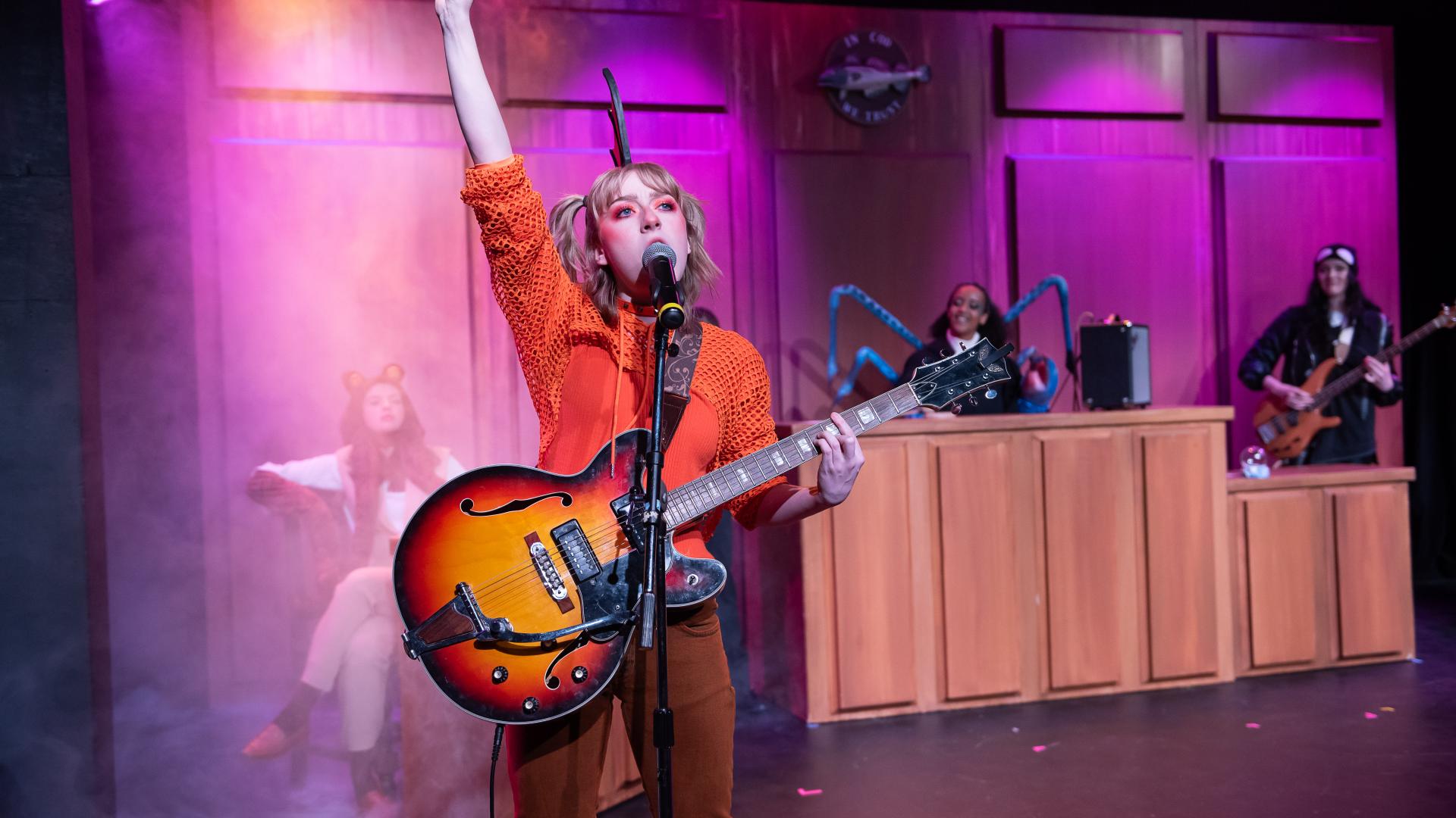 An actor dressed in an orange top with a fin headband plays a guitar at a standing mic. One hand is up in the air in a rockstar-like pose. In the background on stage is a courtroom setup.