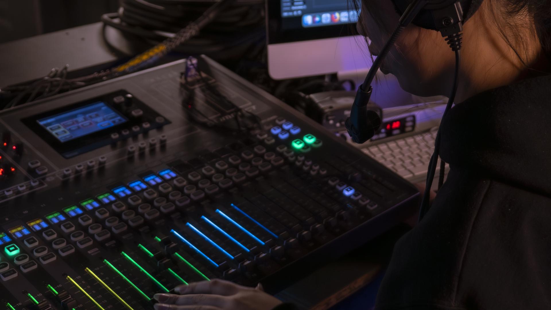 A person sit in front of a technical board glowing green and blue. They are wearing a headset and looking down intently at the board.