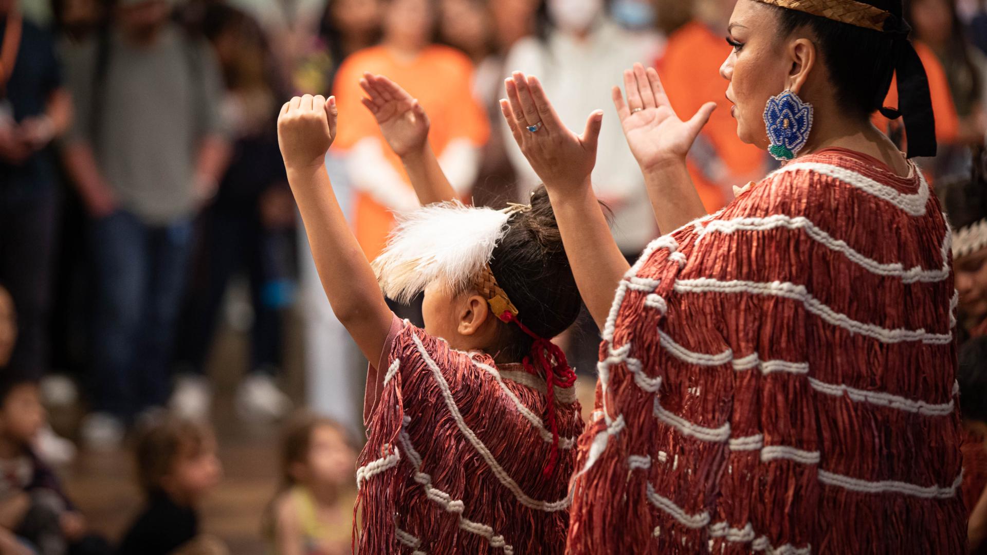 Indigenous woman and child in traditional attire performing with raised arms before a seated audience.