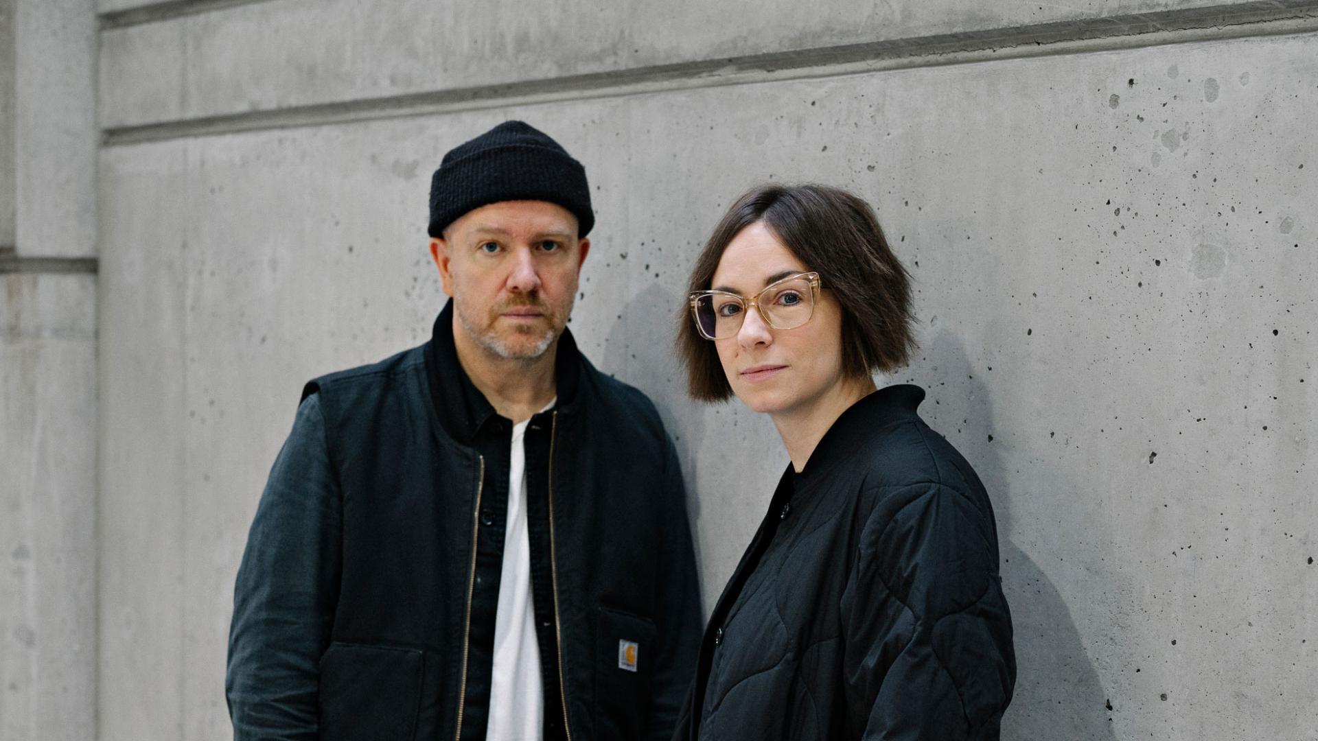Tanya Goehring and Trevor Jacobson (The Automatic Message) posing against a grey wall.