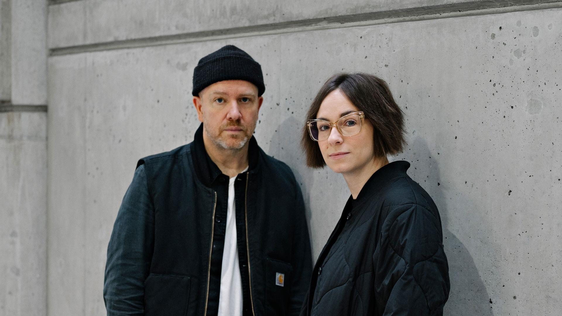 Tanya Goehring and Trevor Jacobson (The Automatic Message) posing against a grey wall.