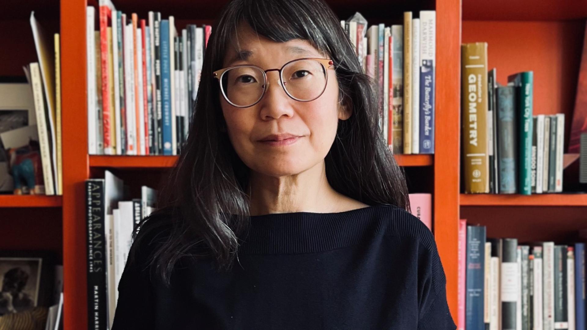 Madeleine Thien portrait standing in front of an orange bookshelf filled with literary works. Photo by Rawi Hage.