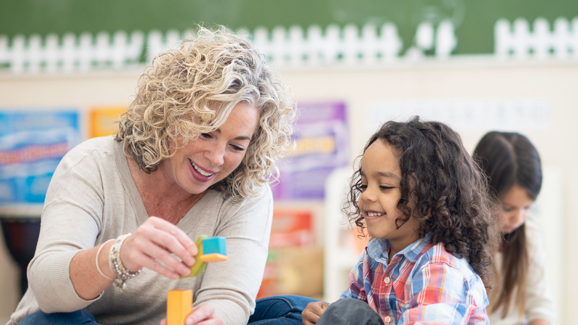 Blonde woman playing blocks with a young student in a classroom setting.