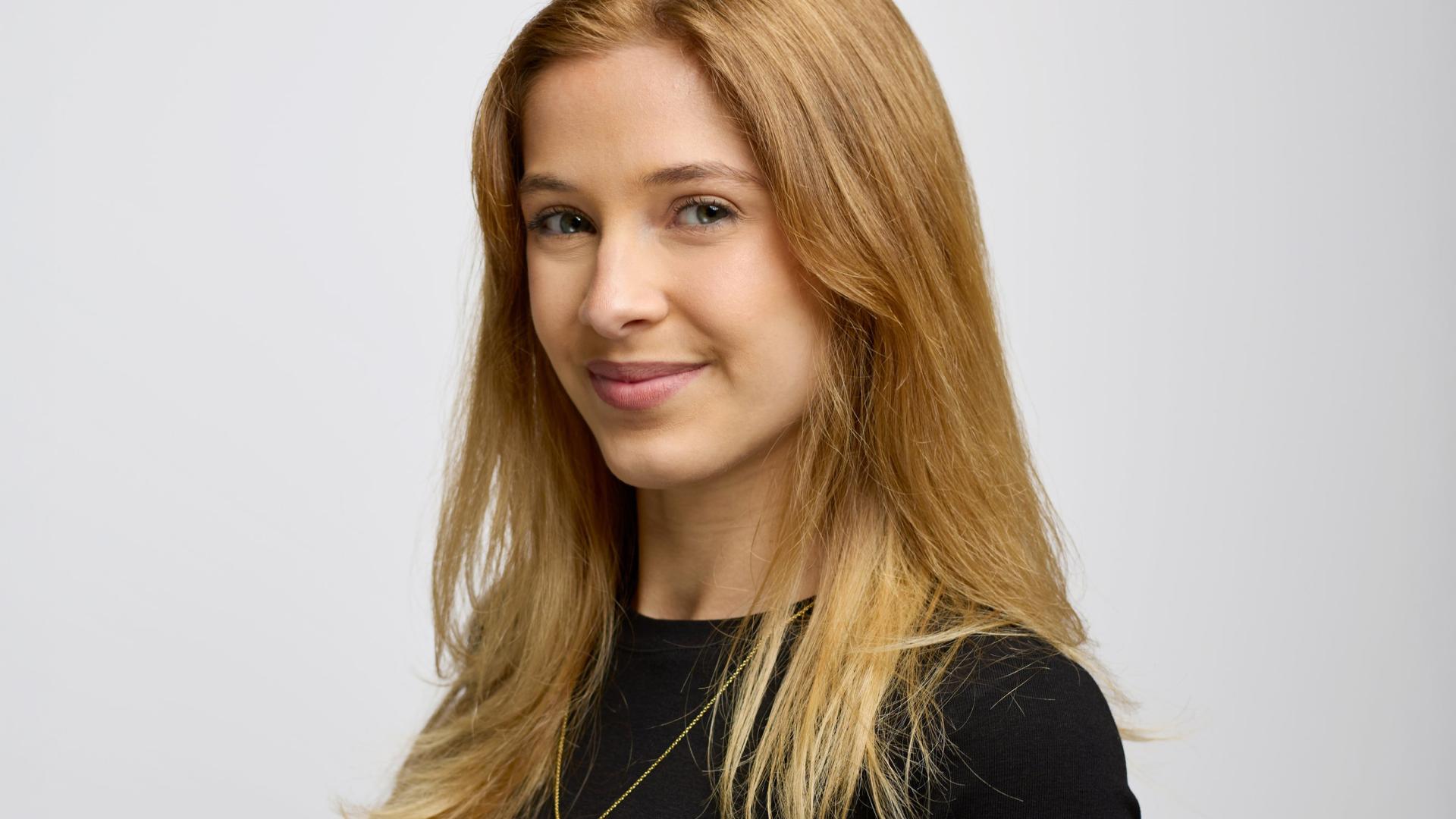 Smiling person with long light brown hair wearing a black top and a delicate necklace, shown in a side-facing portrait against a plain light background.