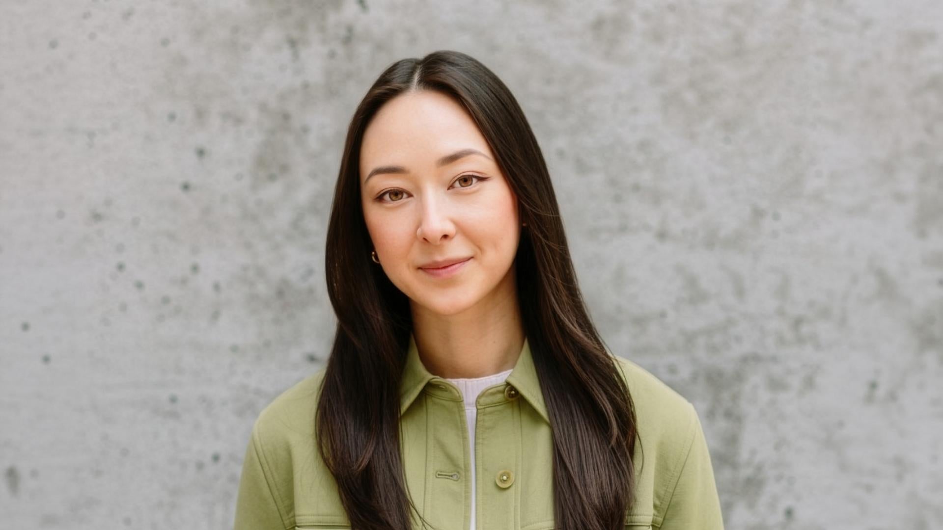 Person with long straight hair wearing a light green jacket and black jeans, standing against a textured gray concrete wall.