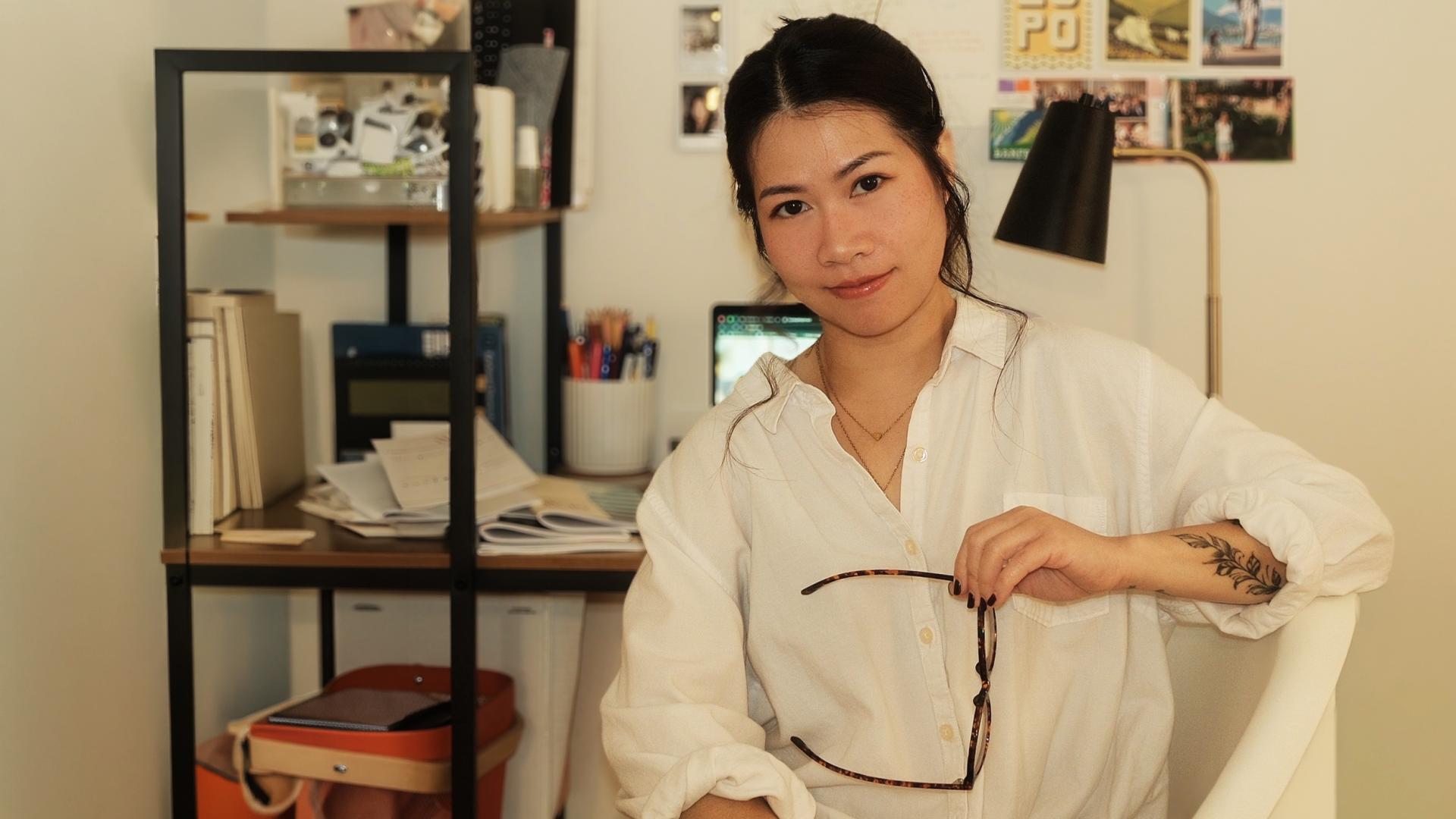 Person sitting in a home office, holding glasses and looking at the camera, with a desk, laptop, books, and photos visible in the background.