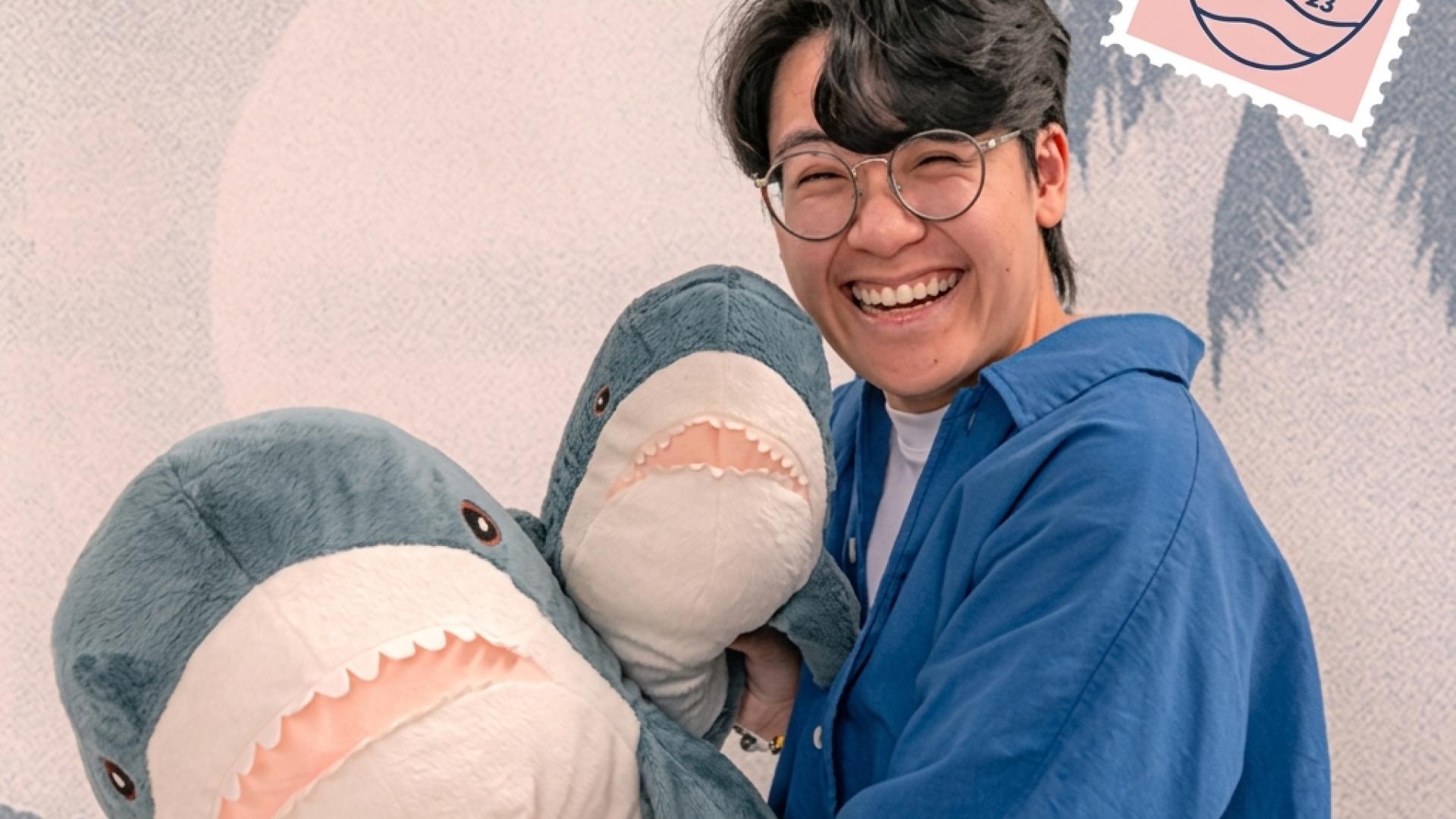 Smiling person wearing glasses and a blue shirt, holding two large plush shark toys in front of a pastel illustration-style background with a small stamp graphic that reads “Illustration 2023.”