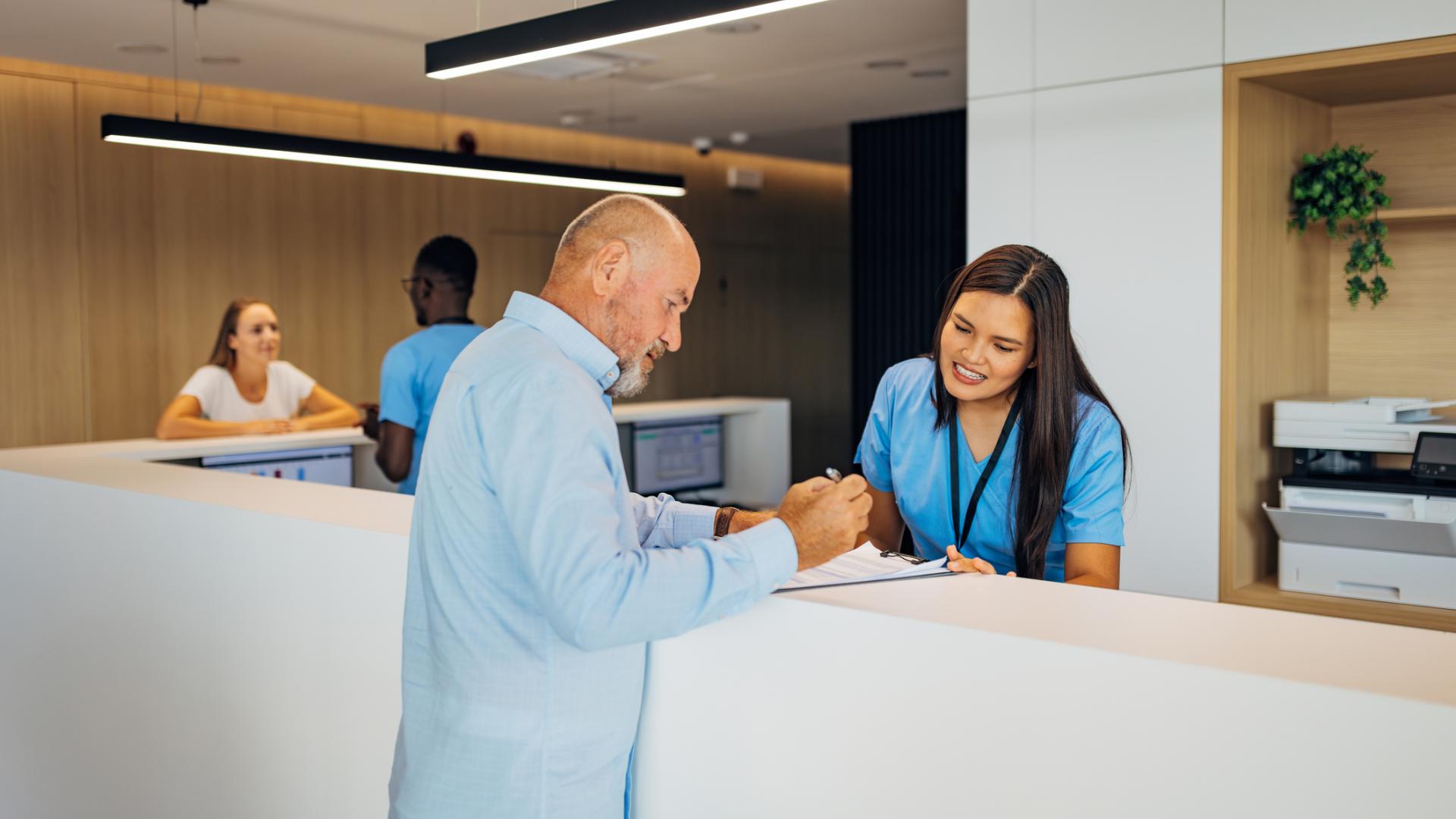 Stock image of the front desk at a medical office, with an office administrator assisting a patient.