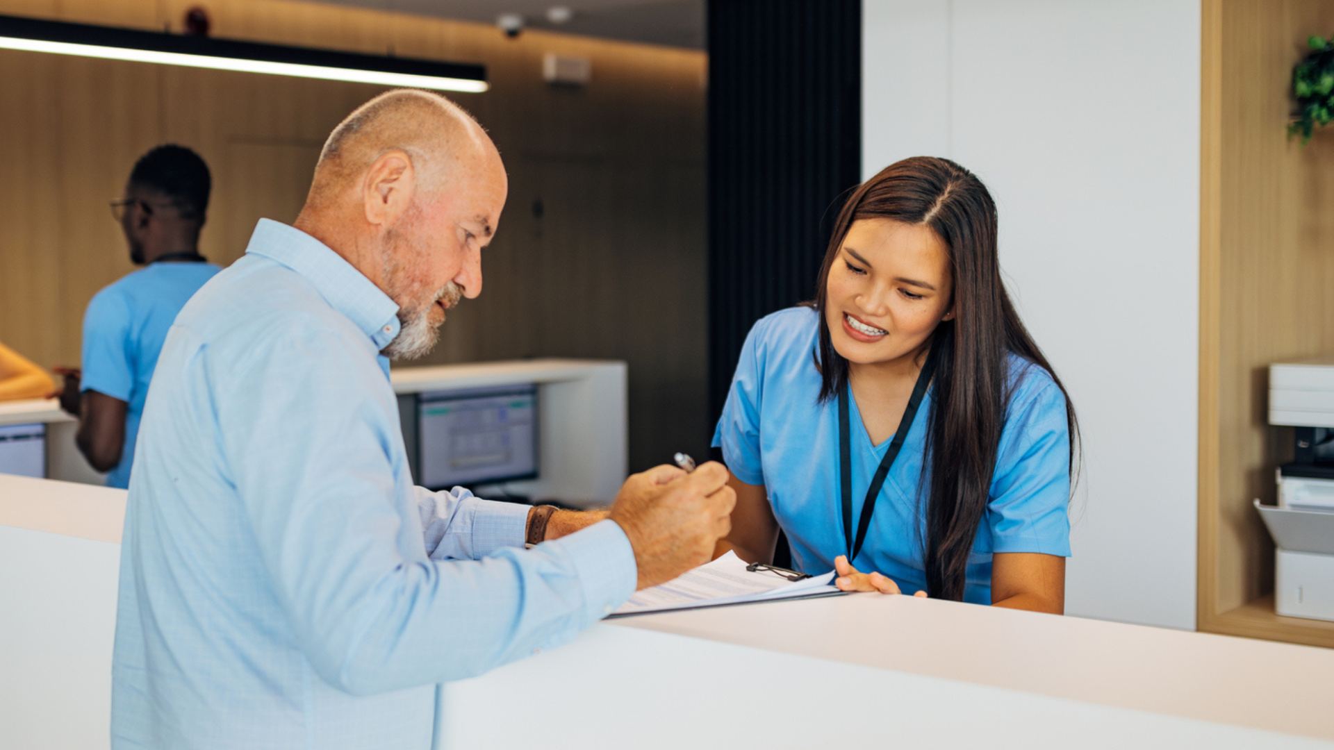 Stock image of the front desk at a medical office, with an office administrator assisting a patient.