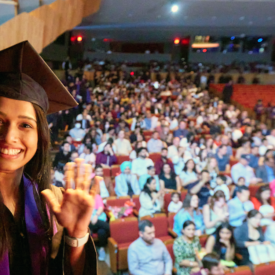 A female student in cap and gown waves and smiles at the camera as she crosses the stage.