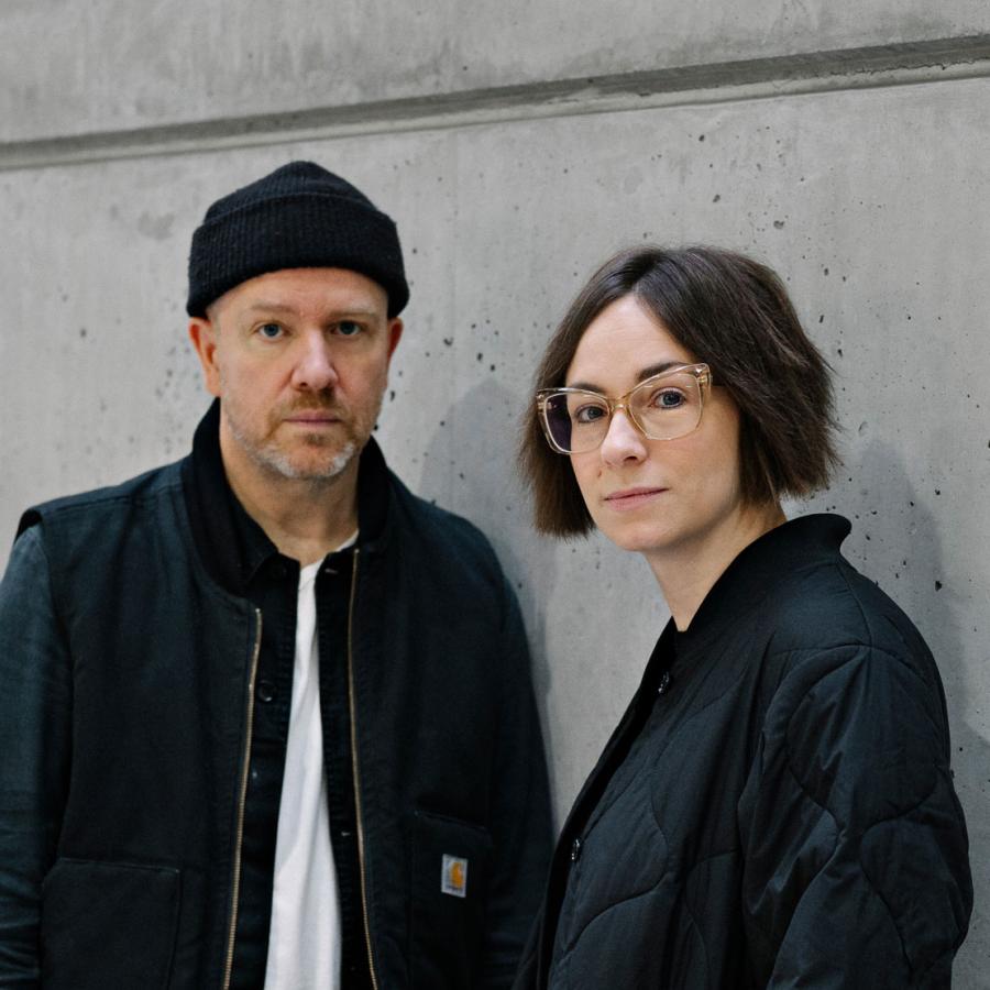 Tanya Goehring and Trevor Jacobson (The Automatic Message) posing against a grey wall.