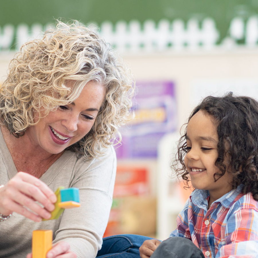 Blonde woman playing blocks with a young student in a classroom setting.