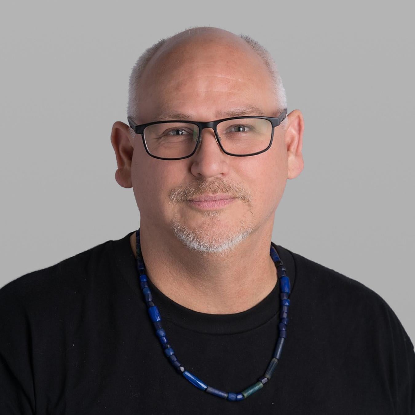 Headshot of Aaron Nelson, with a short grey beard, dark rectangular glasses, a black T-shirt and a blue beaded necklace, against a grey background.