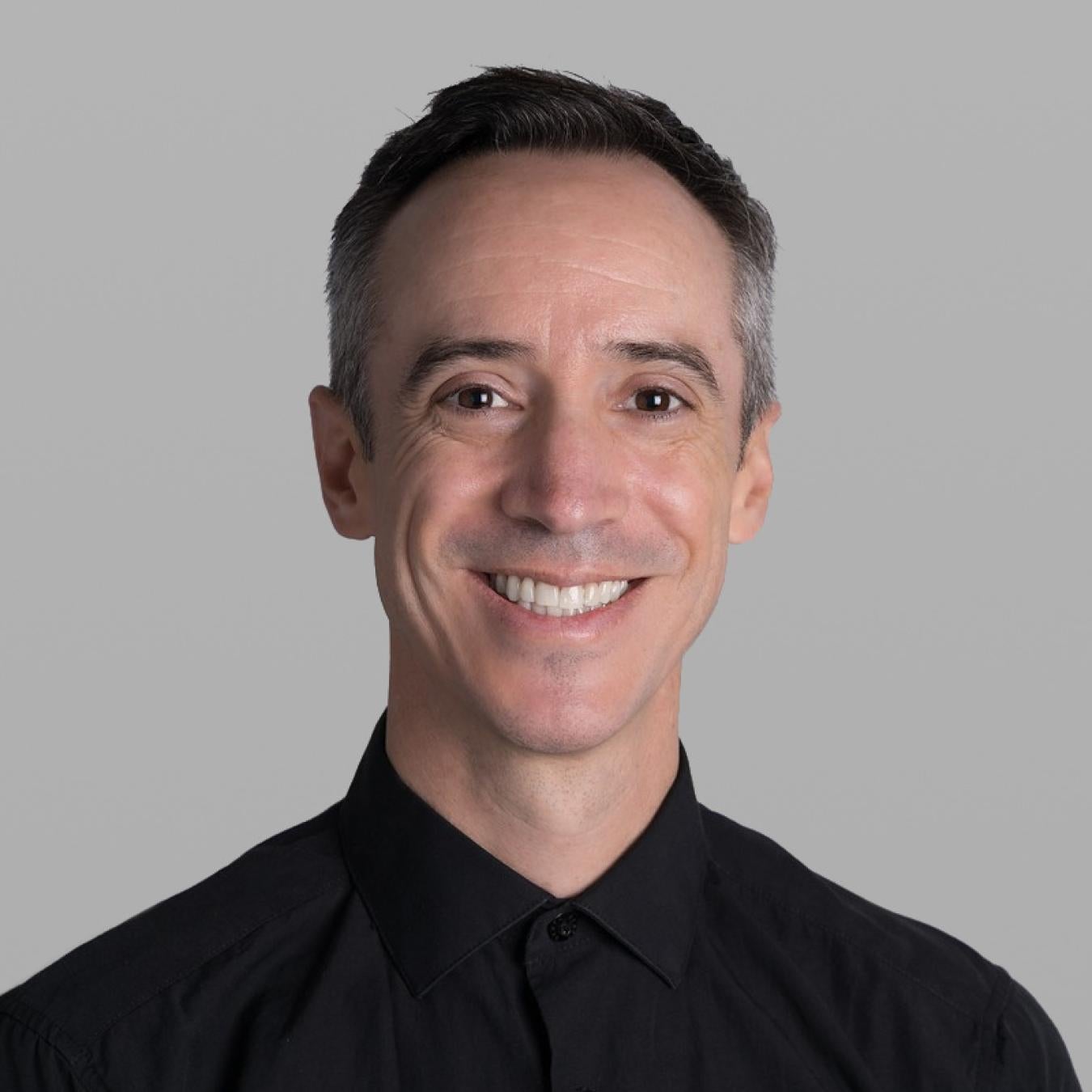 Headshot of Andres Lara Seedorf, smiling, with short greying hair and a black button-up shirt, against a grey background.