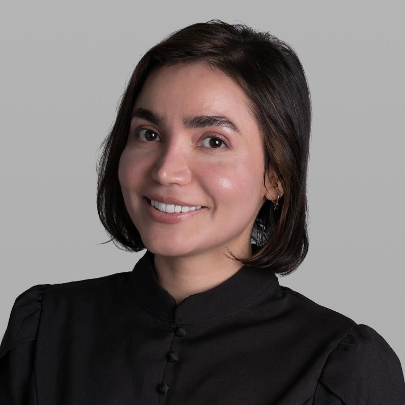 Headshot of Binazir Farokhi, smiling, with a dark chin-length bob and a black high-collared blouse, against a grey background.
