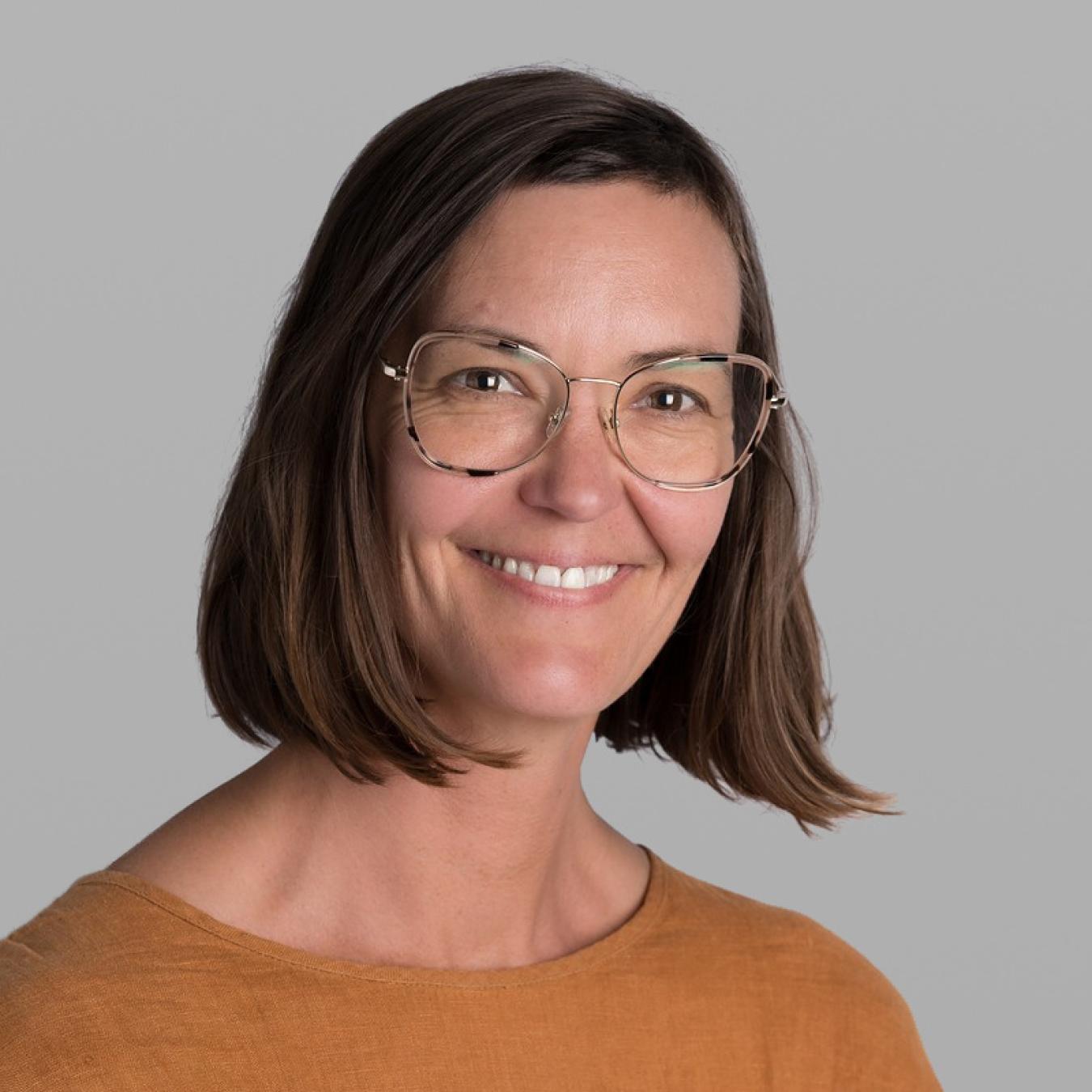 Headshot of Claire Wood, smiling, with shoulder-length brown hair, gold wire-rimmed glasses and a rust-orange sweater, against a grey background.