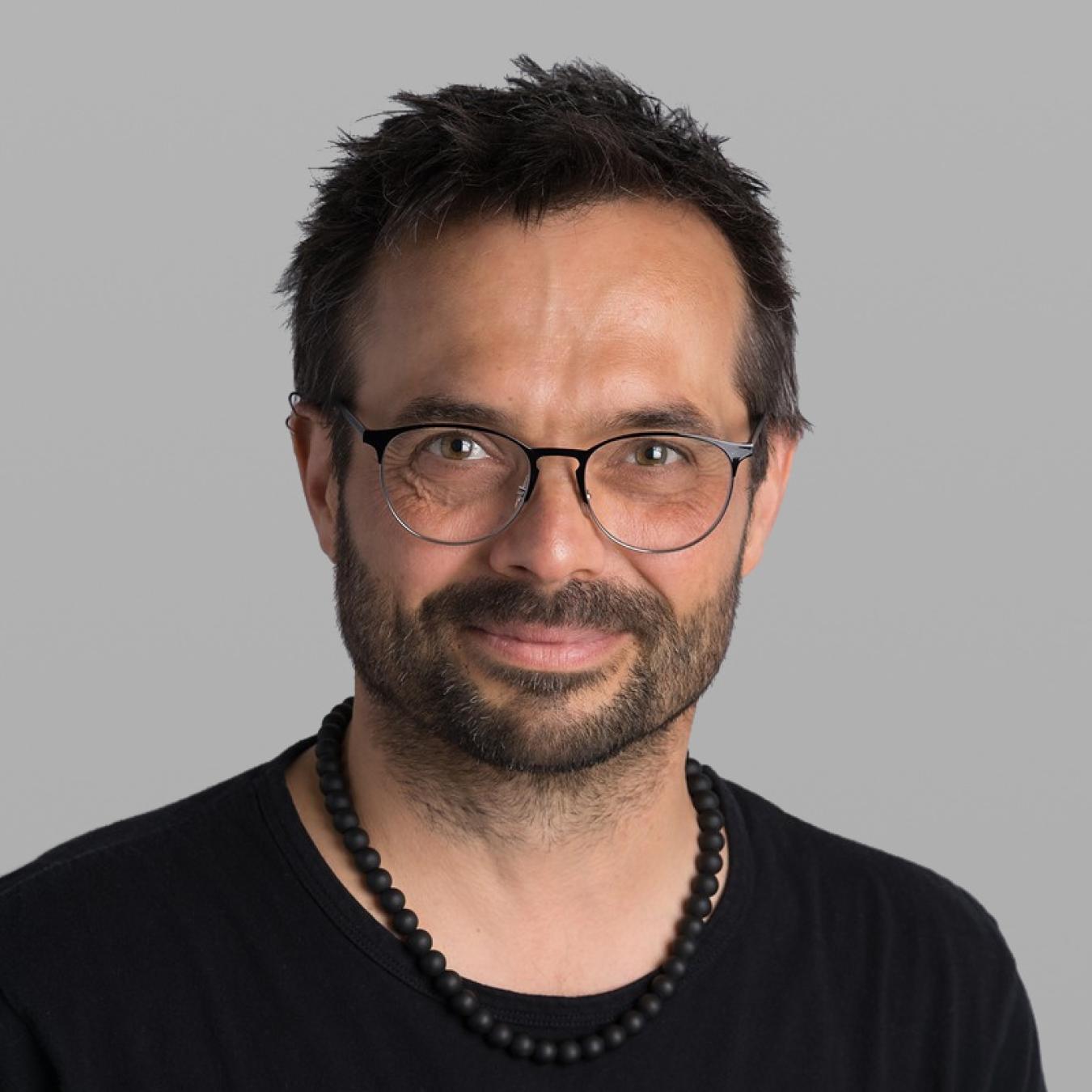 Headshot of Dave Savard, with dark hair, a dark beard, round black glasses, a black T-shirt and a beaded necklace, against a grey background.