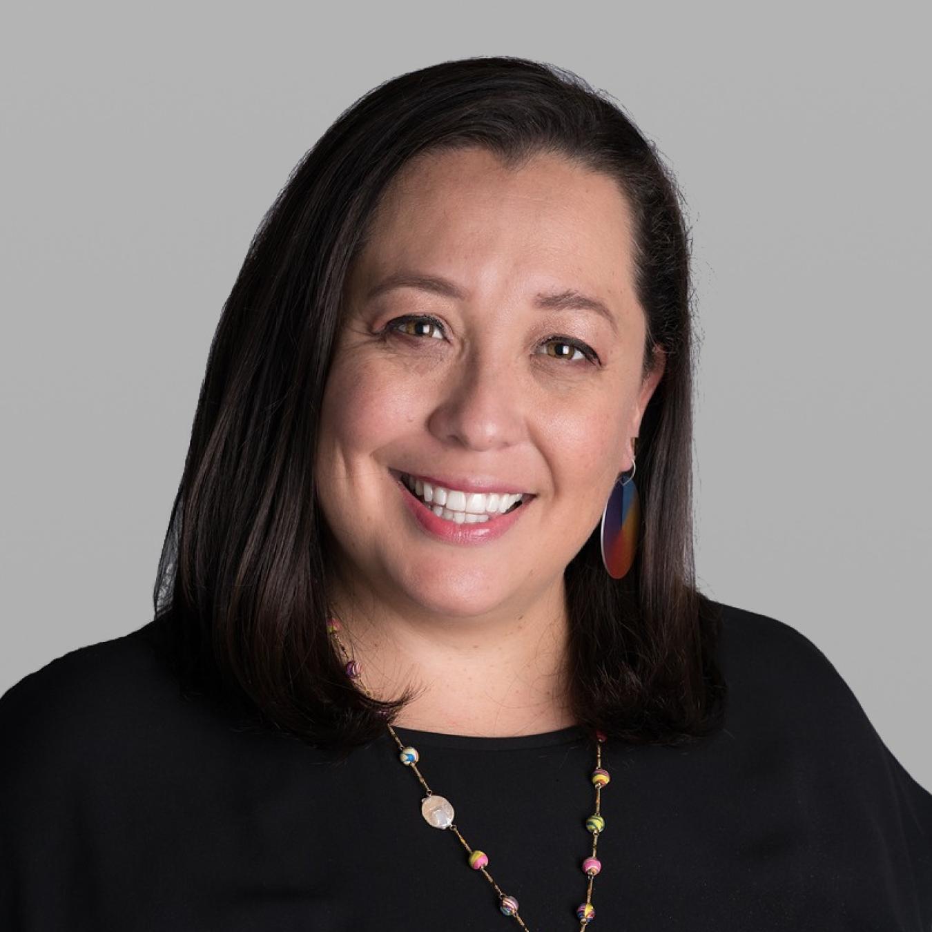 Headshot of Jessica Acevedo Castaneda, smiling, with long dark hair, colourful earrings, a black top and a long beaded necklace, against a grey background.