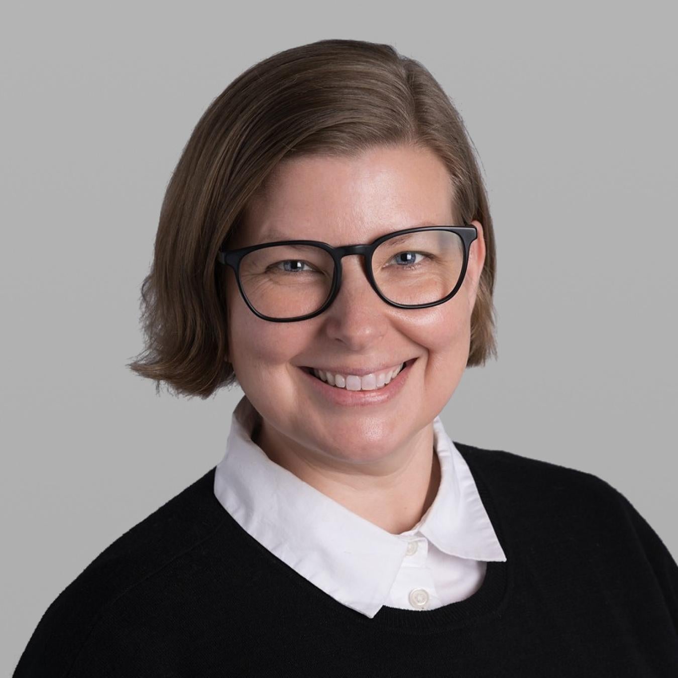 Headshot of Katherine Davis, smiling, with a light brown bob, black glasses and a black sweater over a white collared shirt, against a grey background.