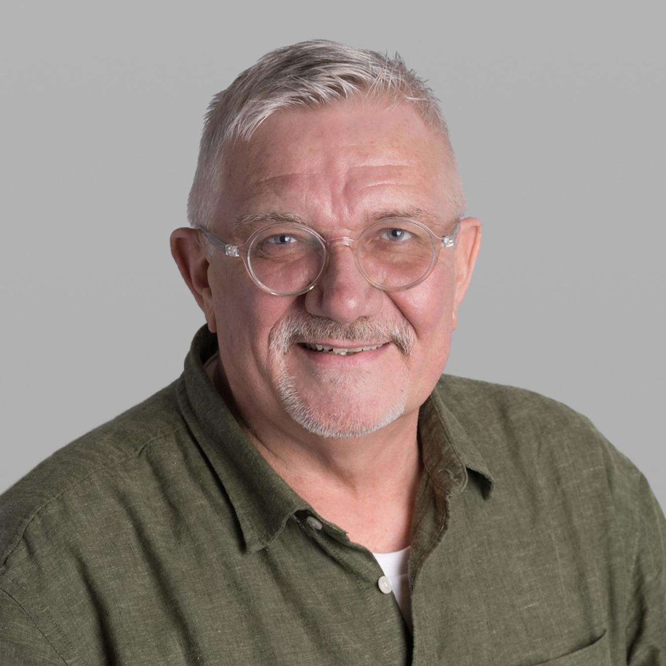 Headshot of Kevin Smith, smiling, with white hair, a short white beard, thin round glasses and an olive-green shirt, against a grey background.