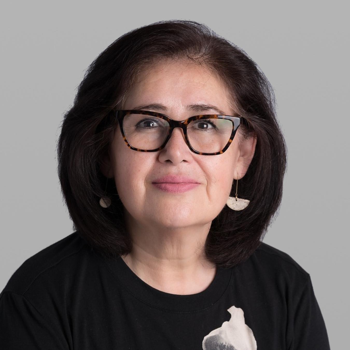Headshot of Marcela Noriega, with dark shoulder-length hair, bold black glasses and a black T-shirt with a white print, against a grey background.
