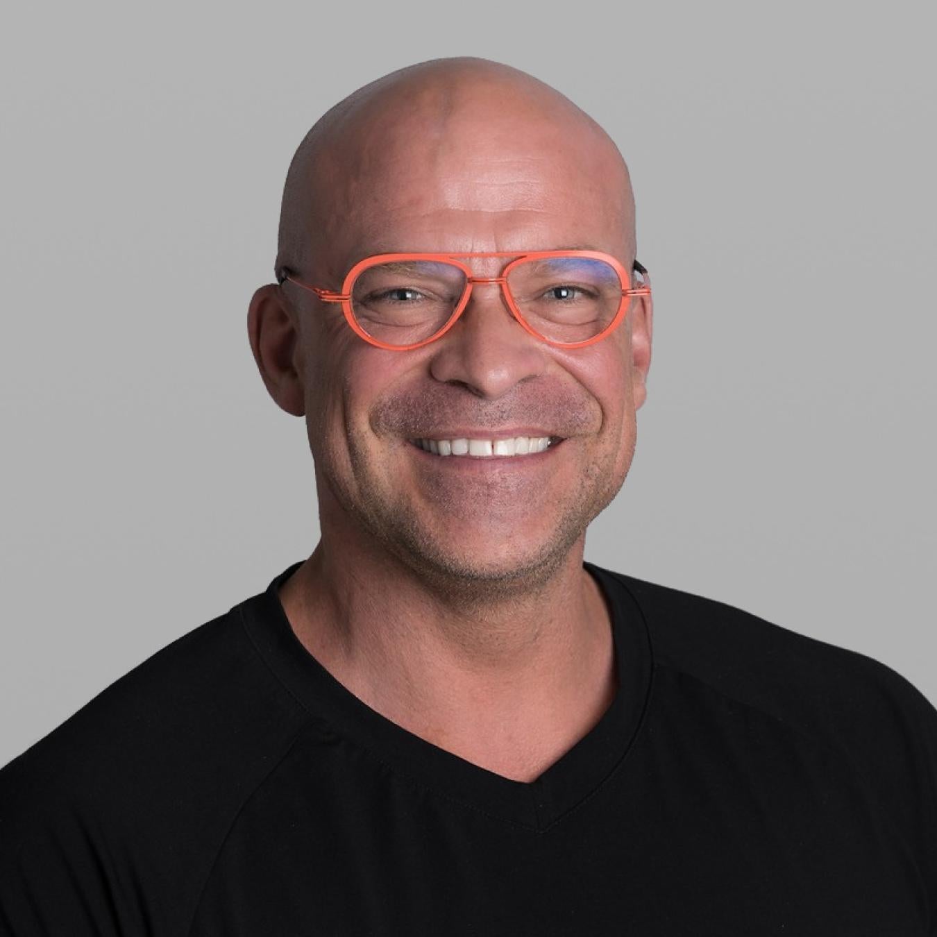 Headshot of Peter Vysek, smiling broadly, with a shaved head, coral-orange glasses and a black V-neck T-shirt, against a grey background.