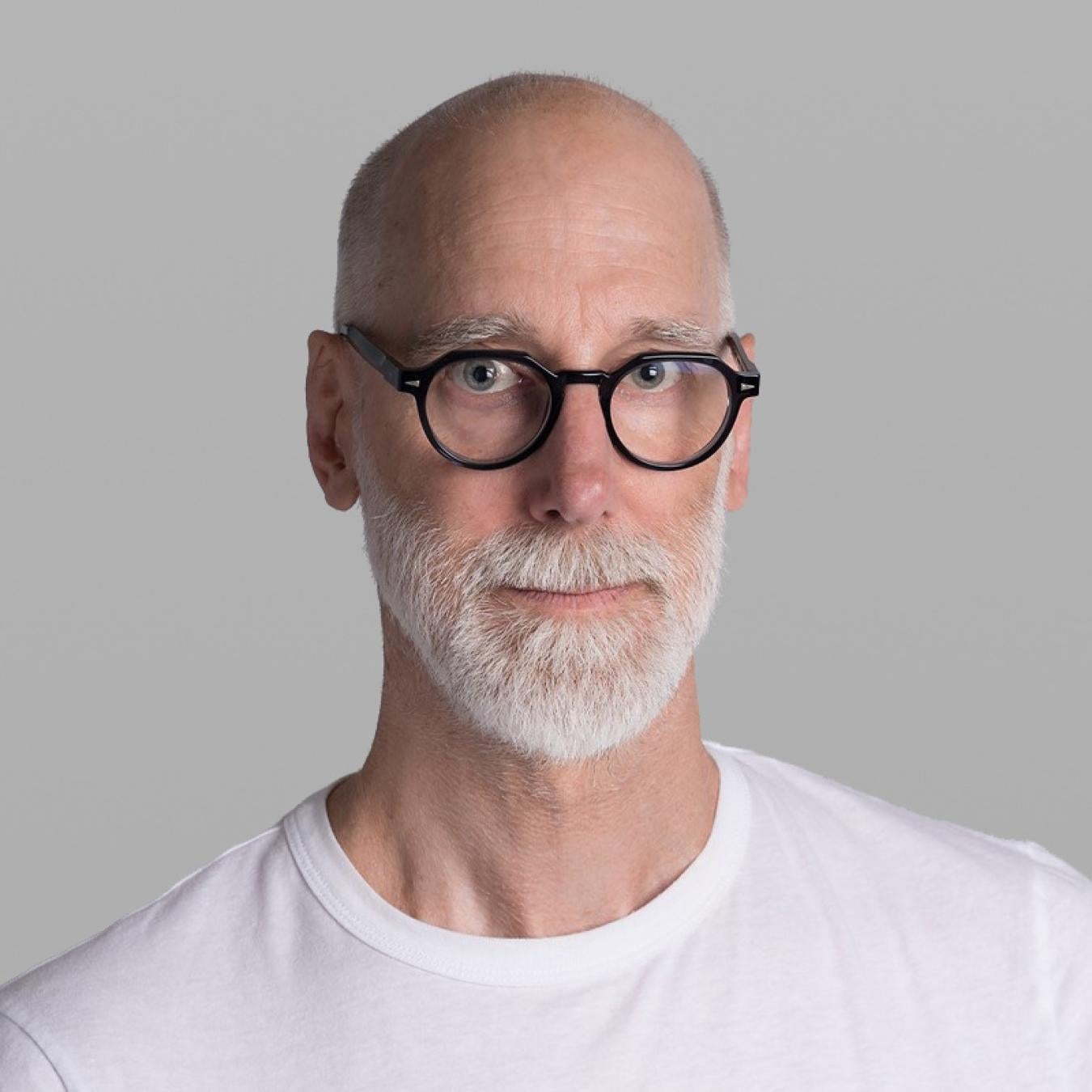 Headshot of Scot Geib, with a shaved head, a white beard, round black glasses and a white T-shirt, against a grey background.