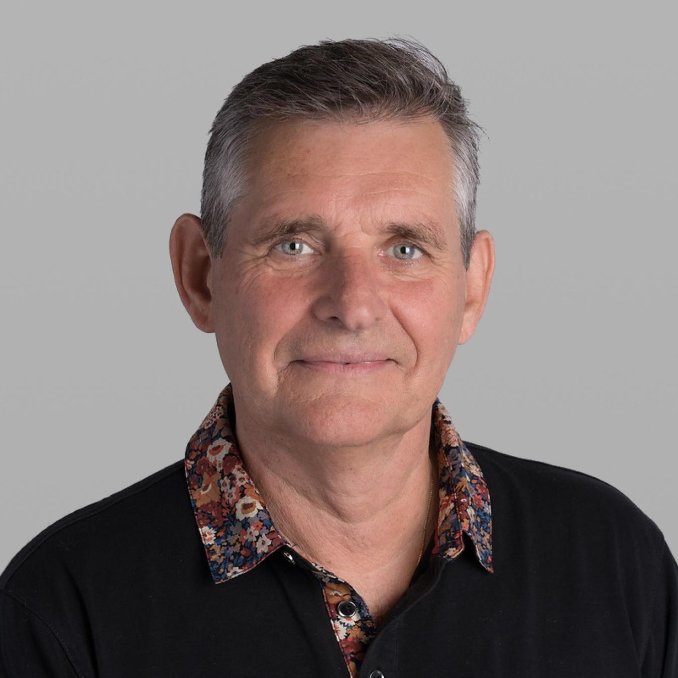 Headshot of Terry Van Roon, with short grey hair and a black jacket over a floral-patterned shirt, against a grey background.