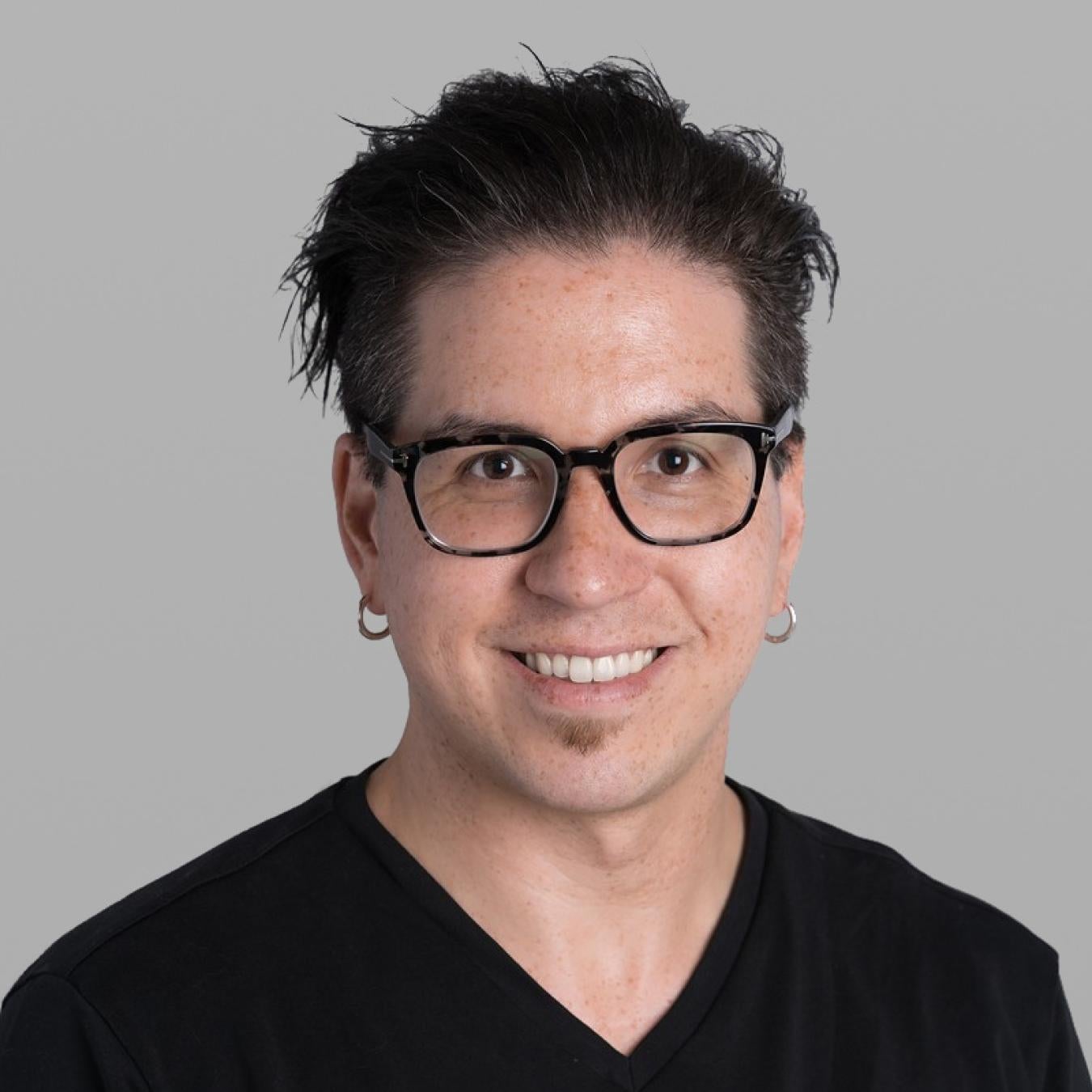 Headshot of Tyler Higgs, smiling, with dark tousled hair, black-framed glasses, small hoop earrings and a black V-neck T-shirt, against a grey background.