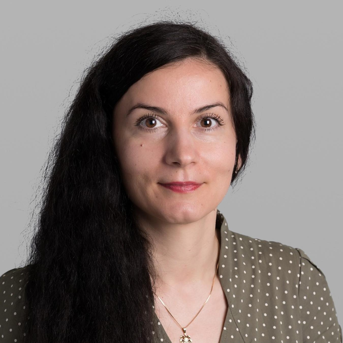 Headshot of Valentina Abanina, with long dark wavy hair, a small pendant necklace and an olive-green polka-dot blouse, against a grey background.