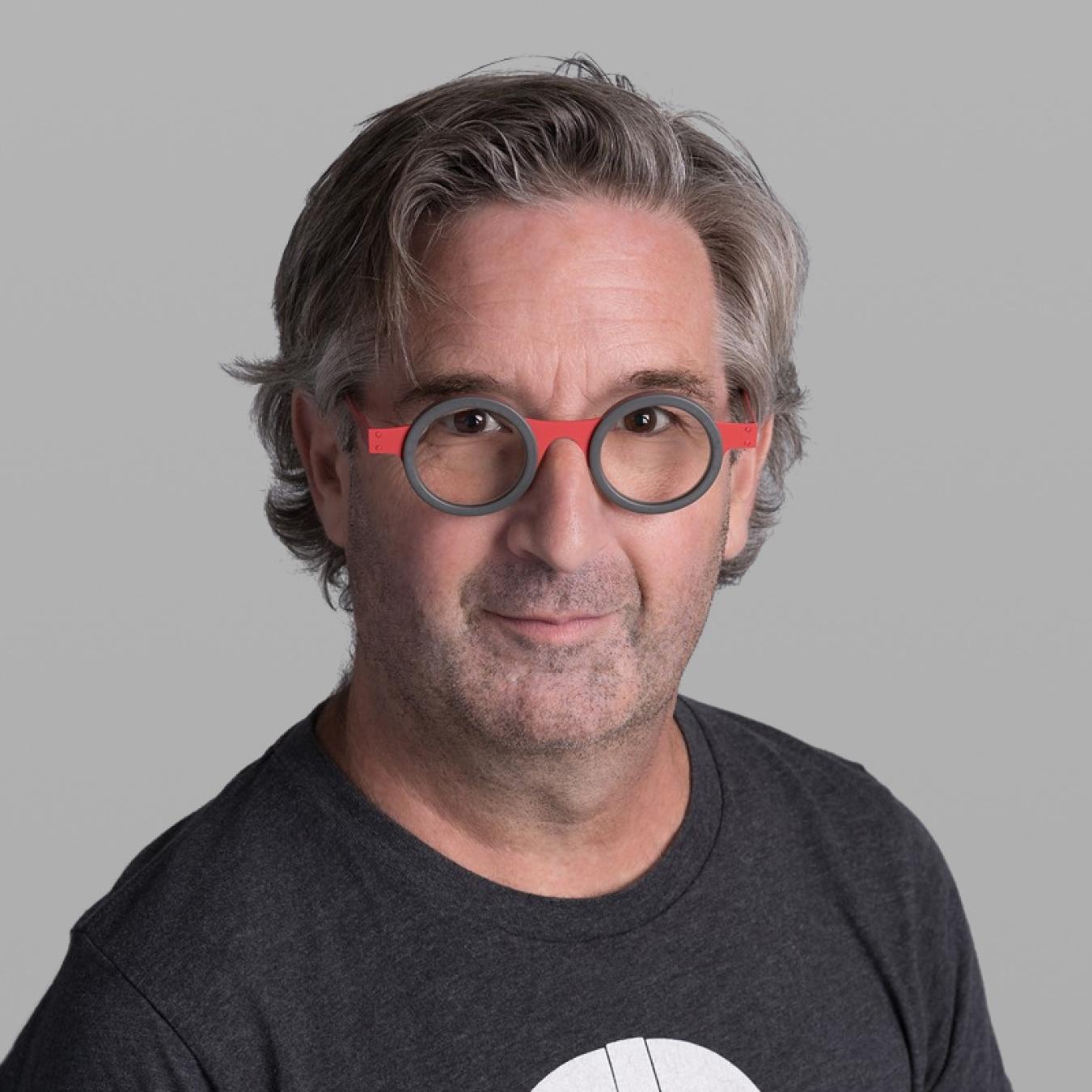 Headshot of Yvan Morissette, with wavy grey hair, round red-and-grey glasses and a dark grey printed T-shirt, against a grey background.