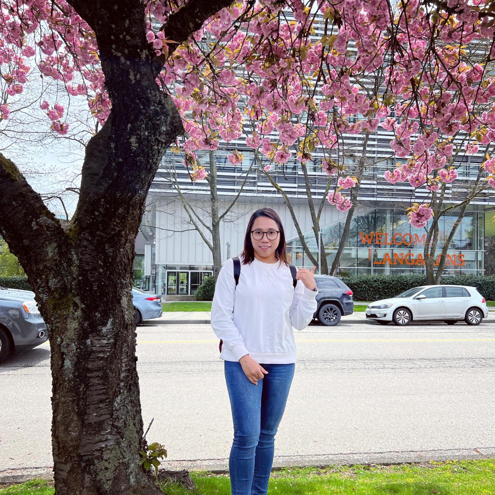 Hnin (Elisa) Pwint under a cherry blossom tree across the street from Langara College with the Science and Technology Building in the background.