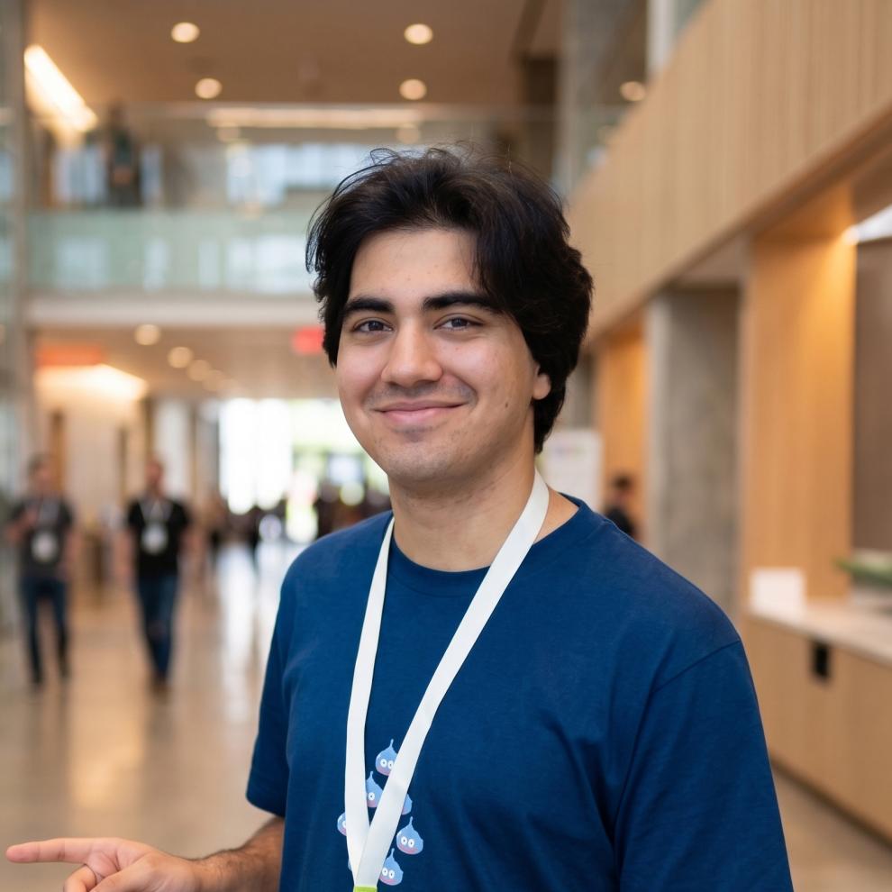 Smiling student with dark, medium-length hair wearing a blue T-shirt and a white lanyard, standing in a bright, modern hallway with people and glass walls in the background.