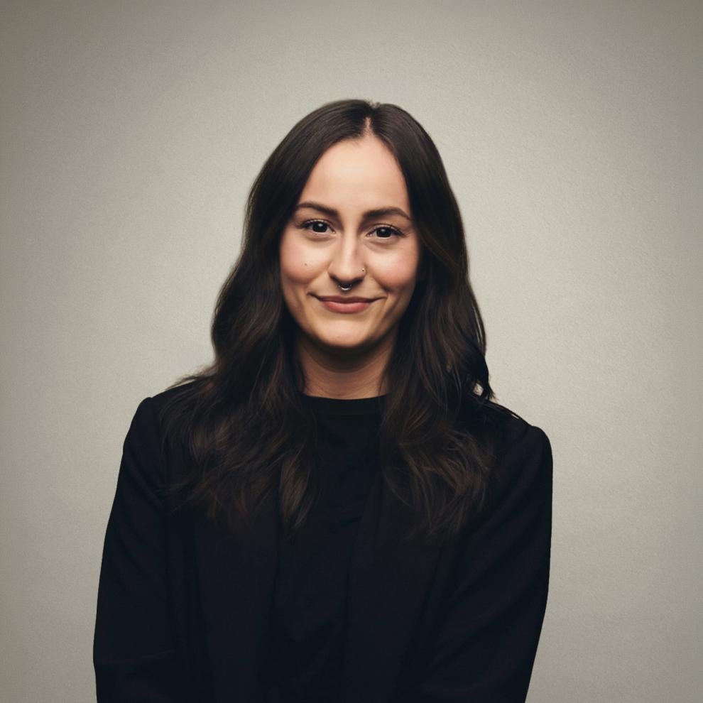 Smiling person with long dark wavy hair and a nose ring, wearing a black top and blazer, posed against a plain light-grey background.