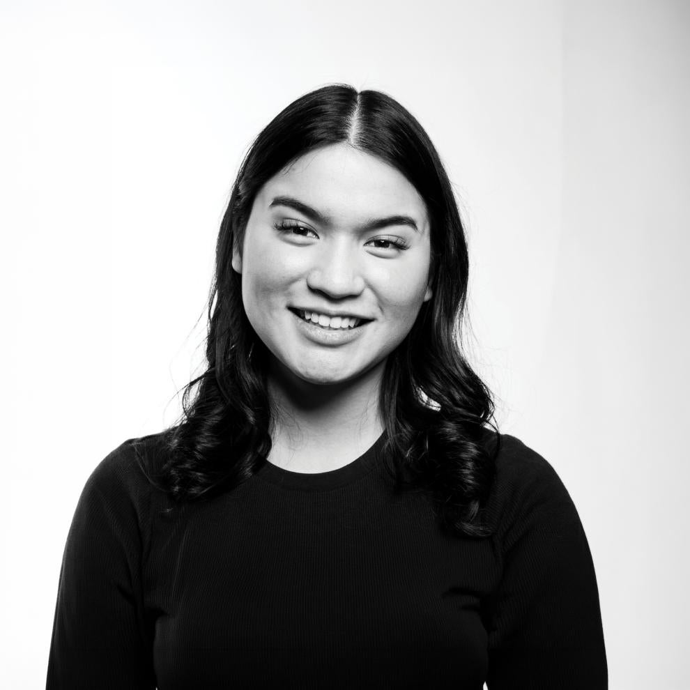 Smiling portrait of a person with shoulder-length dark hair wearing a dark top, shown in black and white against a plain light background.