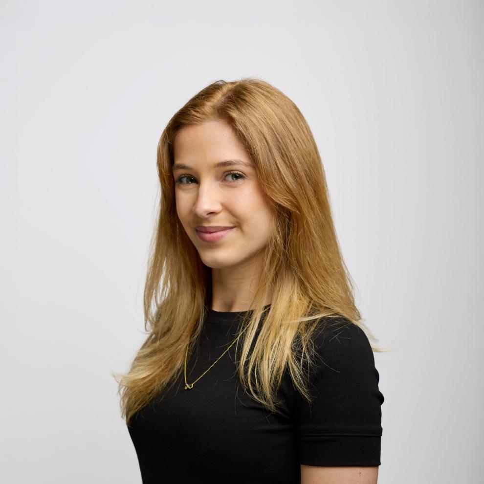 Smiling person with long light brown hair wearing a black top and a delicate necklace, shown in a side-facing portrait against a plain light background.