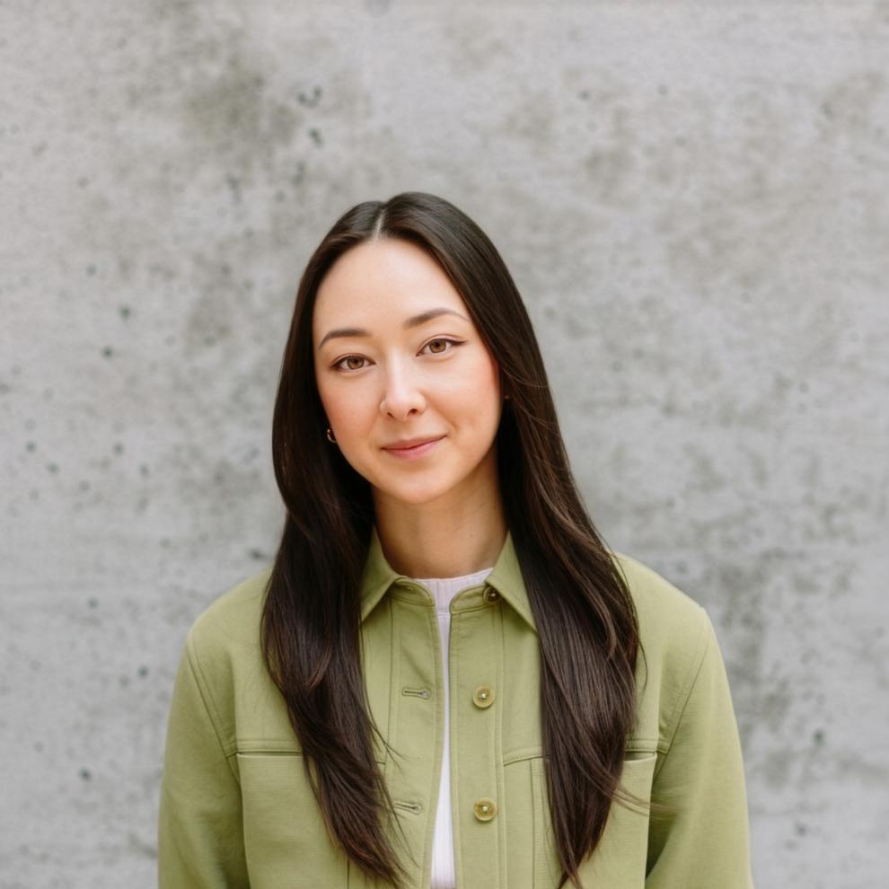 Person with long straight hair wearing a light green jacket and black jeans, standing against a textured gray concrete wall.