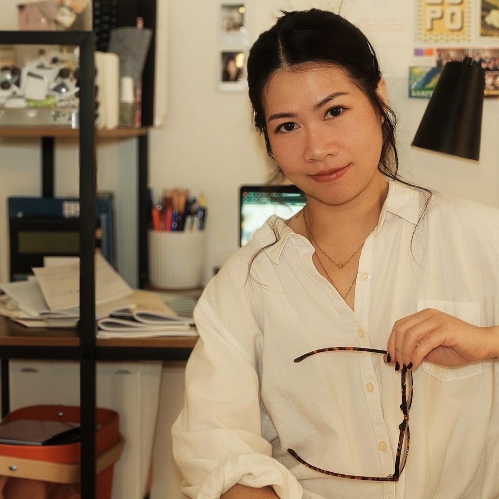 Person sitting in a home office, holding glasses and looking at the camera, with a desk, laptop, books, and photos visible in the background.