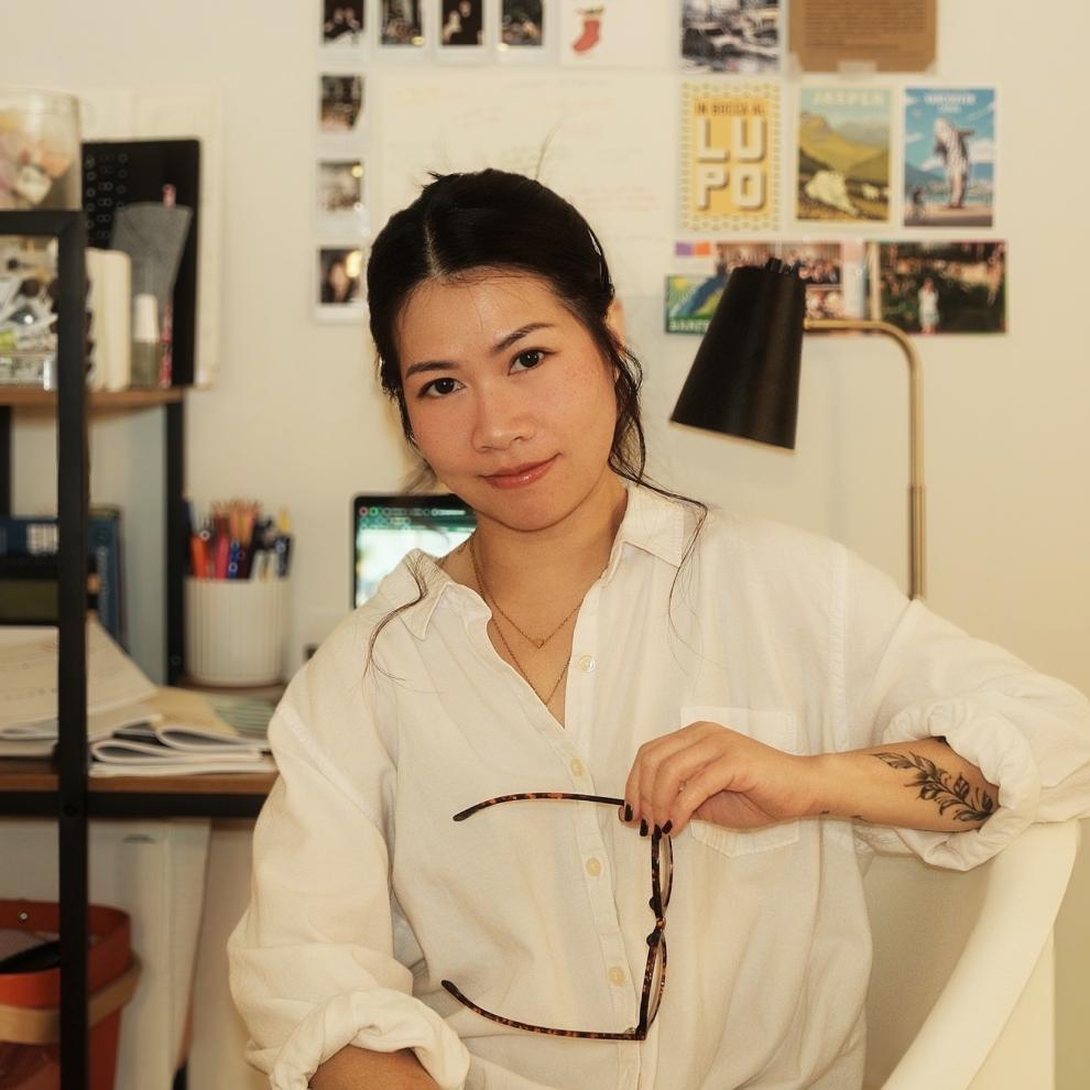 Smiling student wearing a white button-up shirt, holding a pair of glasses while seated in a creative workspace with a desk, lamp, and photo-covered wall in the background.