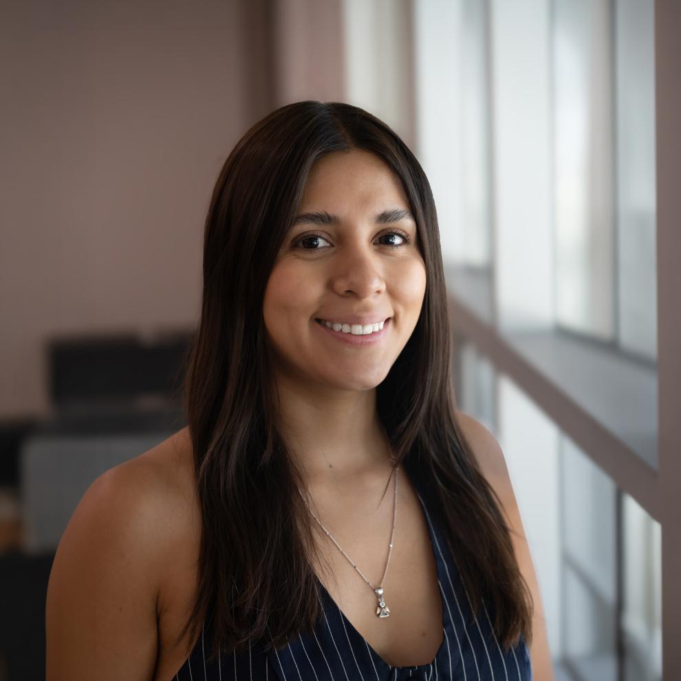 Smiling student with long dark hair wearing a navy pinstriped sleeveless top and a silver necklace, standing indoors beside large windows with a softly blurred background.