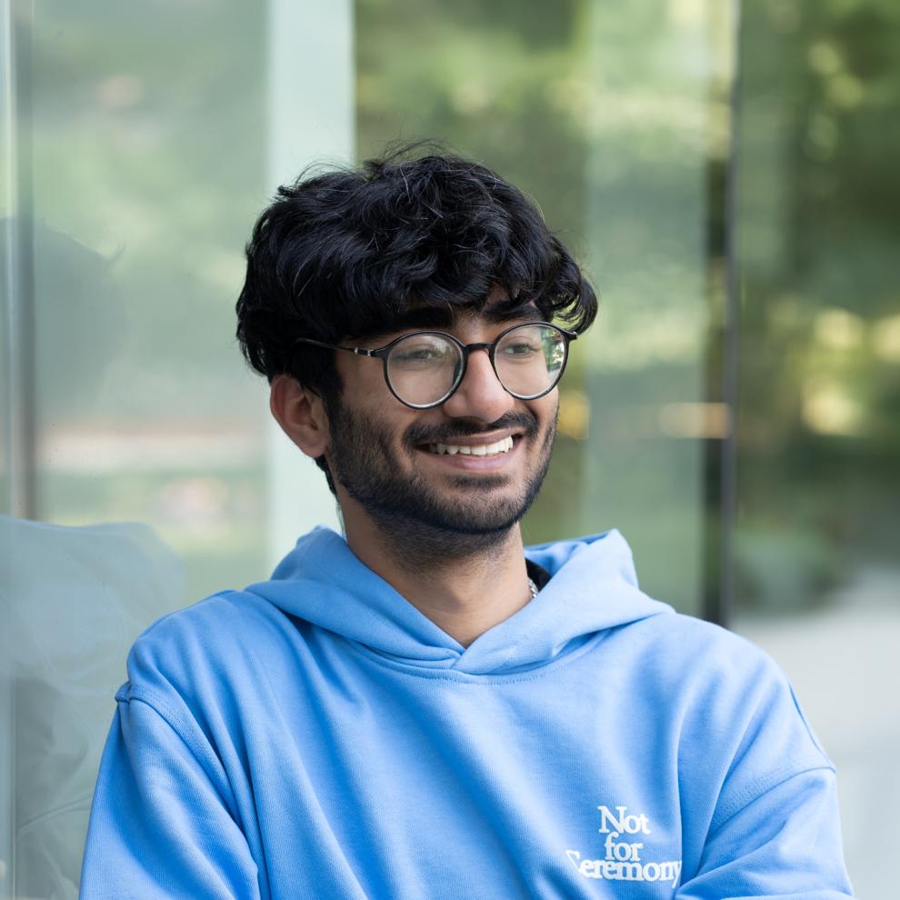 Smiling student wearing round glasses and a light blue hoodie, standing outdoors beside a glass wall with a blurred green background.