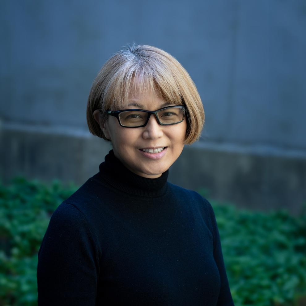 Smiling person with short light brown hair and glasses, wearing a black turtleneck, standing outdoors in front of a blurred blue wall and green foliage.